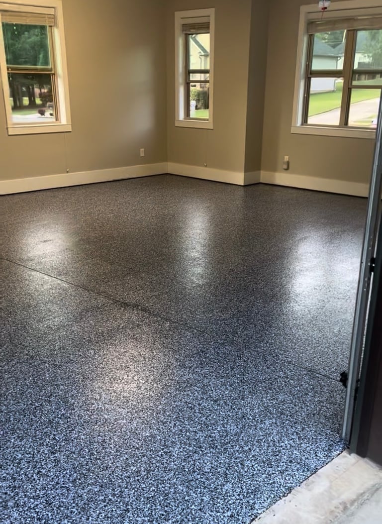 Empty room with beige walls, speckled gray epoxy floor, and three windows overlooking a green yard