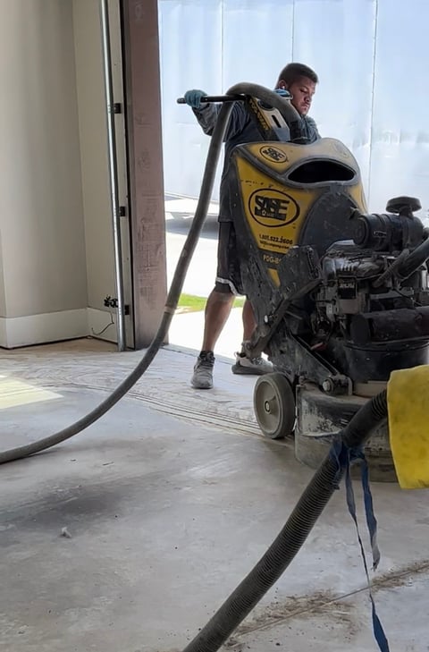 Worker operating yellow SAE concrete grinding machine on indoor floor with large hose