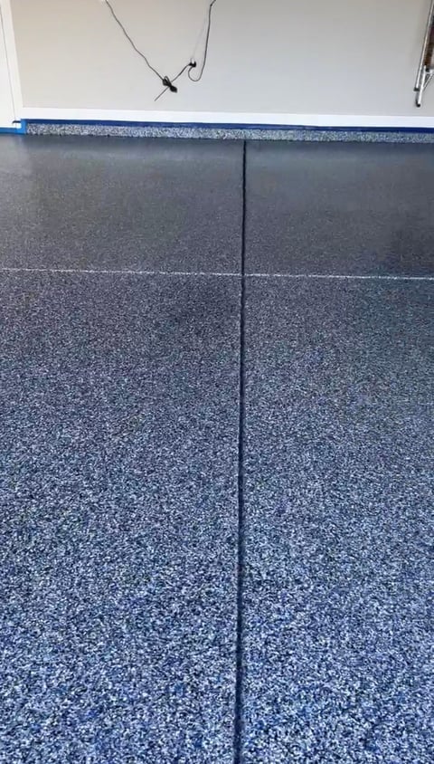 Blue and gray epoxy garage floor with seams between sections and electrical outlet visible on white wall above