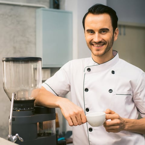 Male chef in white double-breasted chefs jacket smiling while operating a blender in a modern kitchen