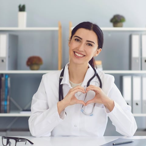Female doctor in white coat and stethoscope making heart shape with hands at office desk