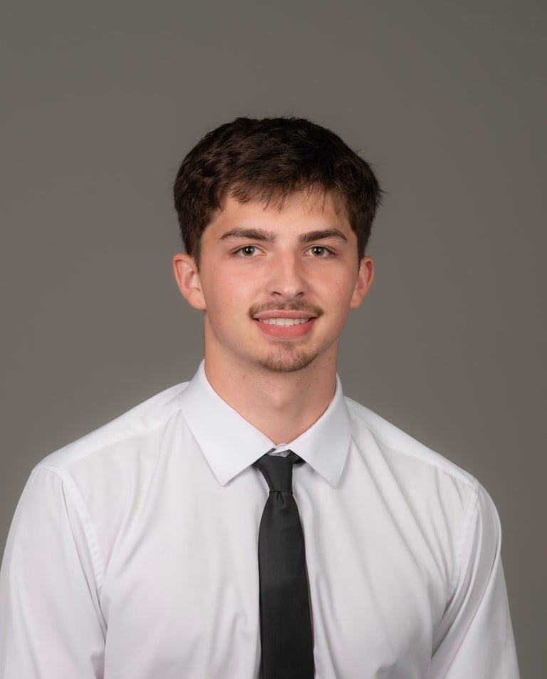 Young man with dark hair and facial hair smiling at camera, wearing white dress shirt and black tie against gray background