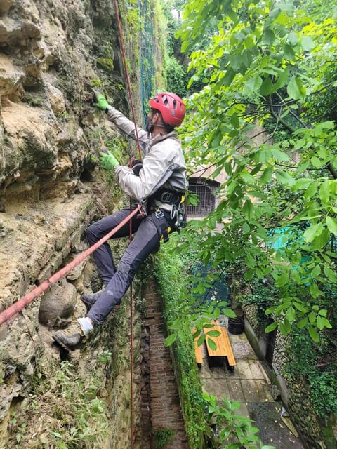 A person in a red helmet and harness rock climbing on an outdoor stone wall surrounded by green vegetation