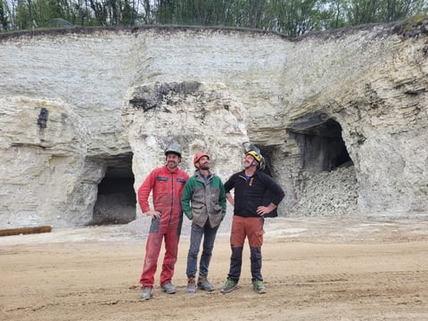 Three people in winter clothing stand on sand in front of a large cliff face with dark cave openings