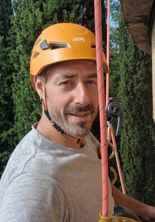 Man wearing yellow climbing helmet and safety gear on rope, smiling at camera outdoors with trees in background