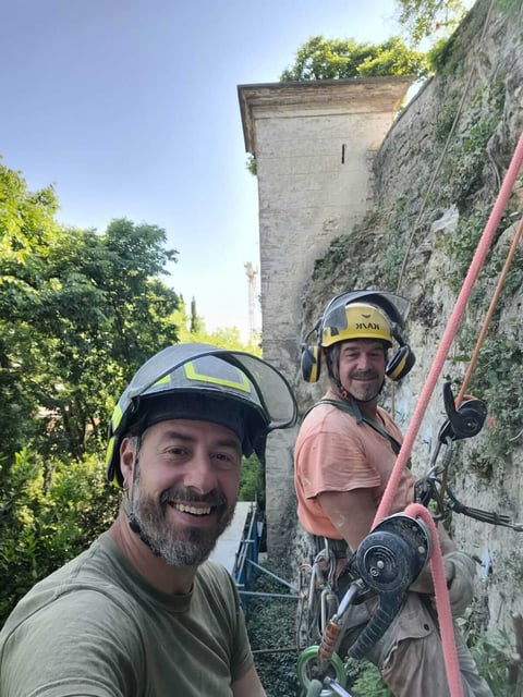 Two rock climbers smile at camera while ascending a stone tower with ivy-covered walls and pink climbing rope
