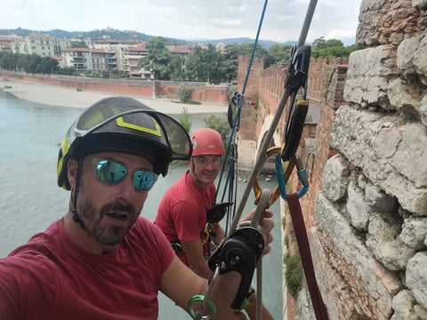 Two men in safety gear parasailing from a rocky cliff overlooking a turquoise river with buildings and hills in the background