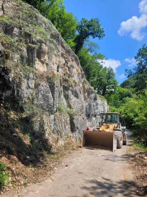 Heavy machinery grading a dirt road alongside a tall stone wall with vegetation, surrounded by lush green trees and blue sky.