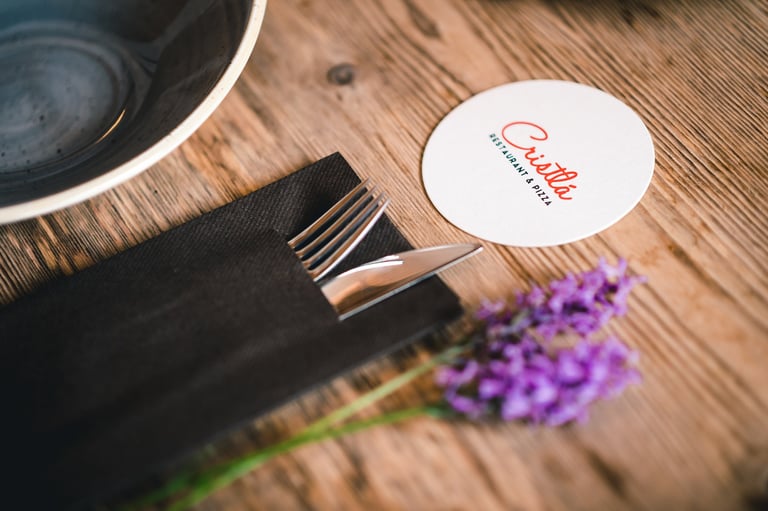 Festlich gedeckter Tisch mit Cristlá-Untersetzer, schwarzer Serviette, Besteck und lila Blumen