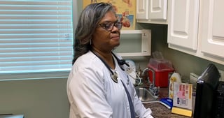 Woman in white lab coat and glasses standing in a medical office or clinic room with cabinets and medical equipment