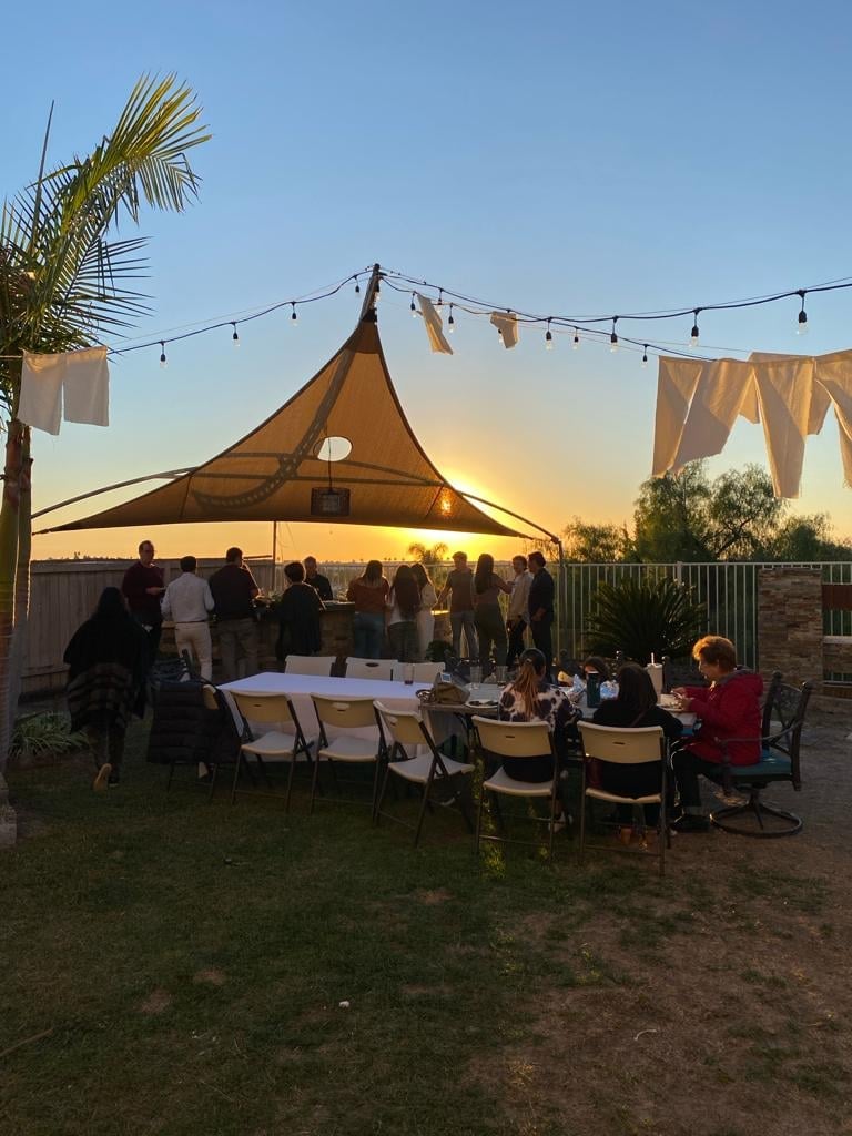 Reunión nocturna al aire libre bajo una carpa color arena con luces, asistentes sentados en mesas y de pie, fondo de atardecer con palmera y telas colgadas