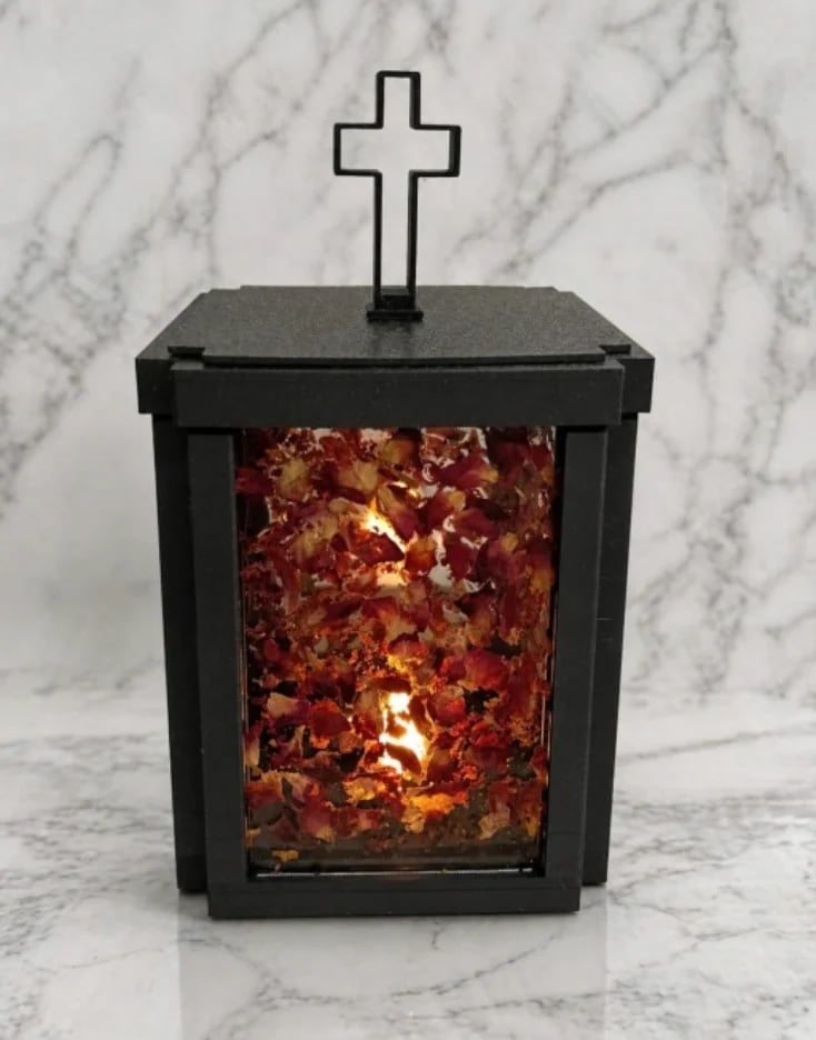 Black cremation urn with white cross on top and red-gold cremains visible through glass window on marble background.