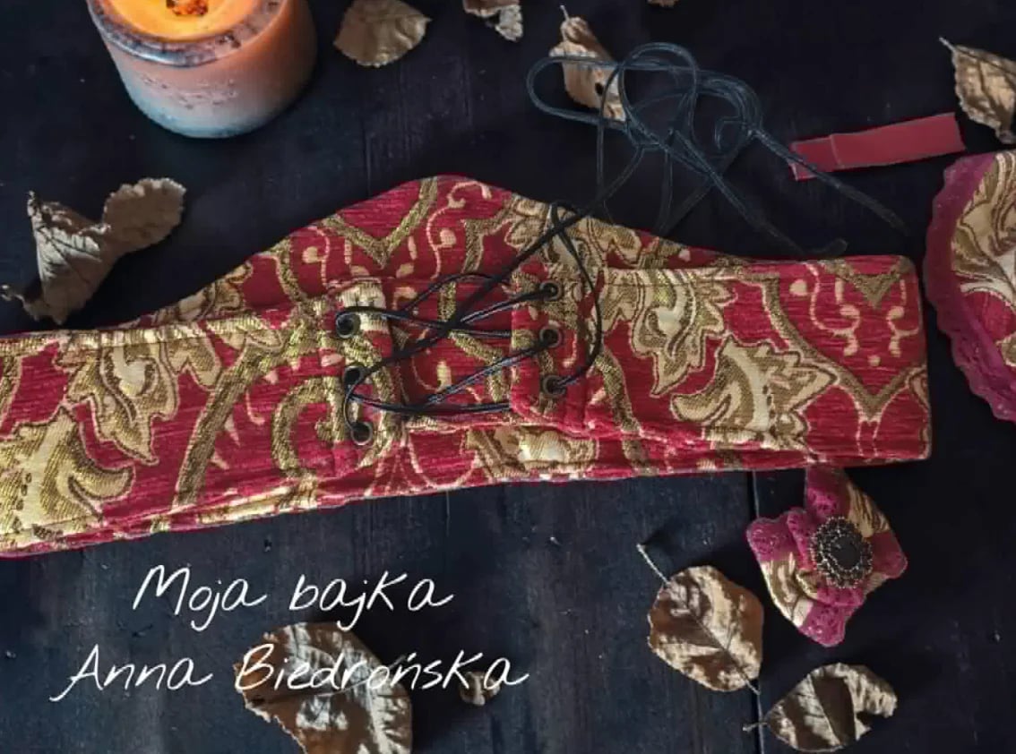 Red and gold embroidered traditional textile piece displayed on dark background with dried botanicals and text reading Moja bajka Anna Biedrzanska