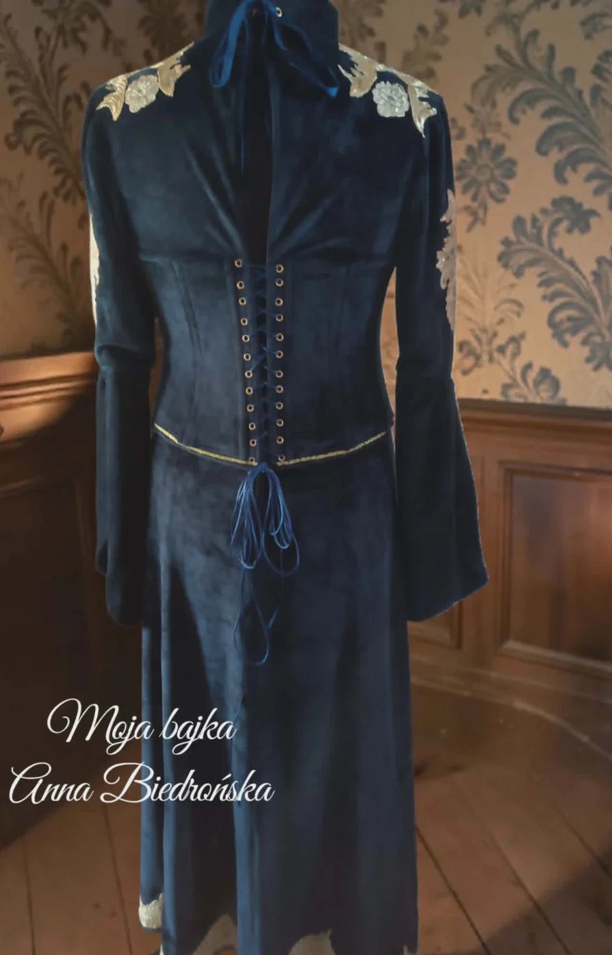 Navy blue medieval-style dress with lace shoulders, corset-style lacing, and flowing sleeves displayed on a mannequin against damask wallpaper