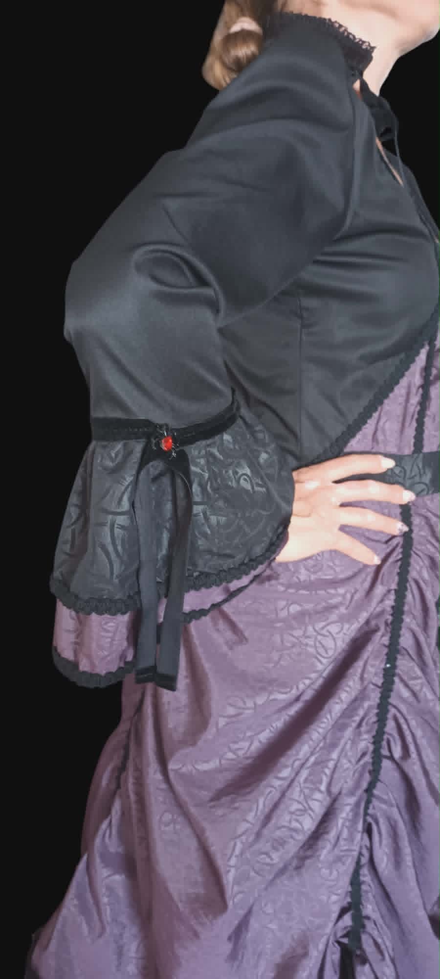 Woman wearing black shrug over purple brocade dress with black corset belt featuring red gem detail