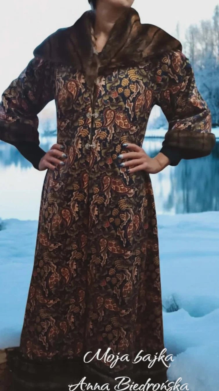 Woman wearing ornate dark dress with gold embroidery standing in snowy landscape with frozen lake background