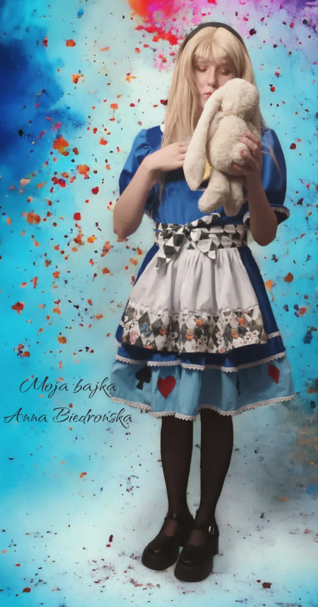 Young girl with blonde hair wearing a blue dress and holding a stuffed bunny against a colorful paint splatter background