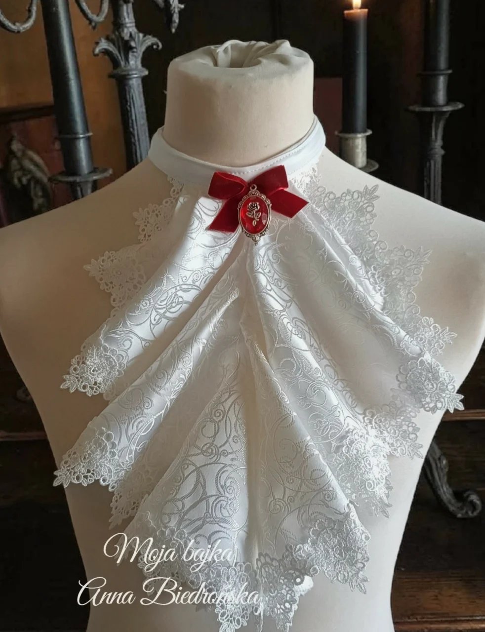 White lace christening cape on dress form with red bow and decorative ornament against dark background