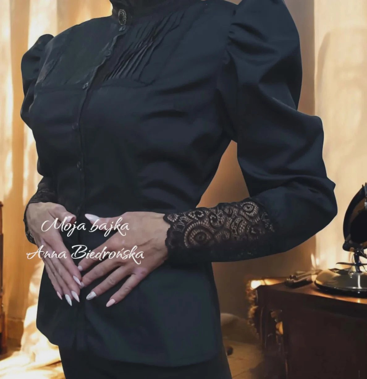 Woman wearing a black dress with puffed sleeves and lace details on the arms, posed against a warm-lit interior background