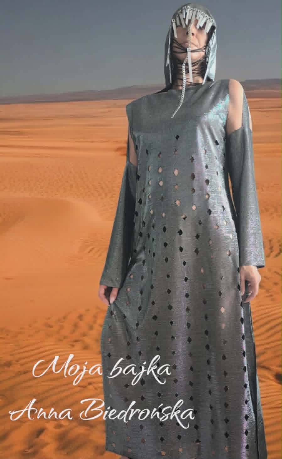 Person wearing a gray perforated dress and metallic head covering standing in a desert landscape with sand dunes