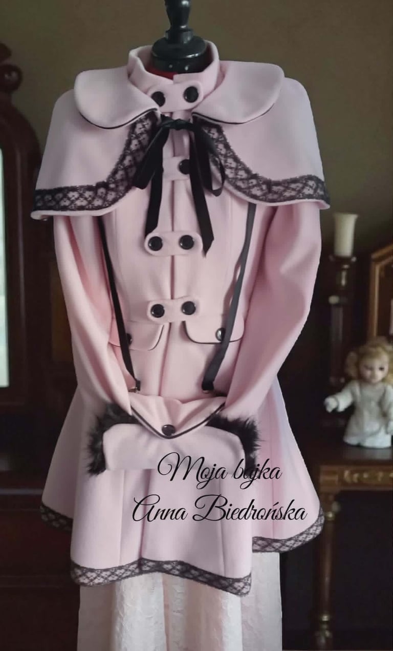 Pink and white mannequin dress with black lace trim, cape collar, and black bow details, displaying text in Polish