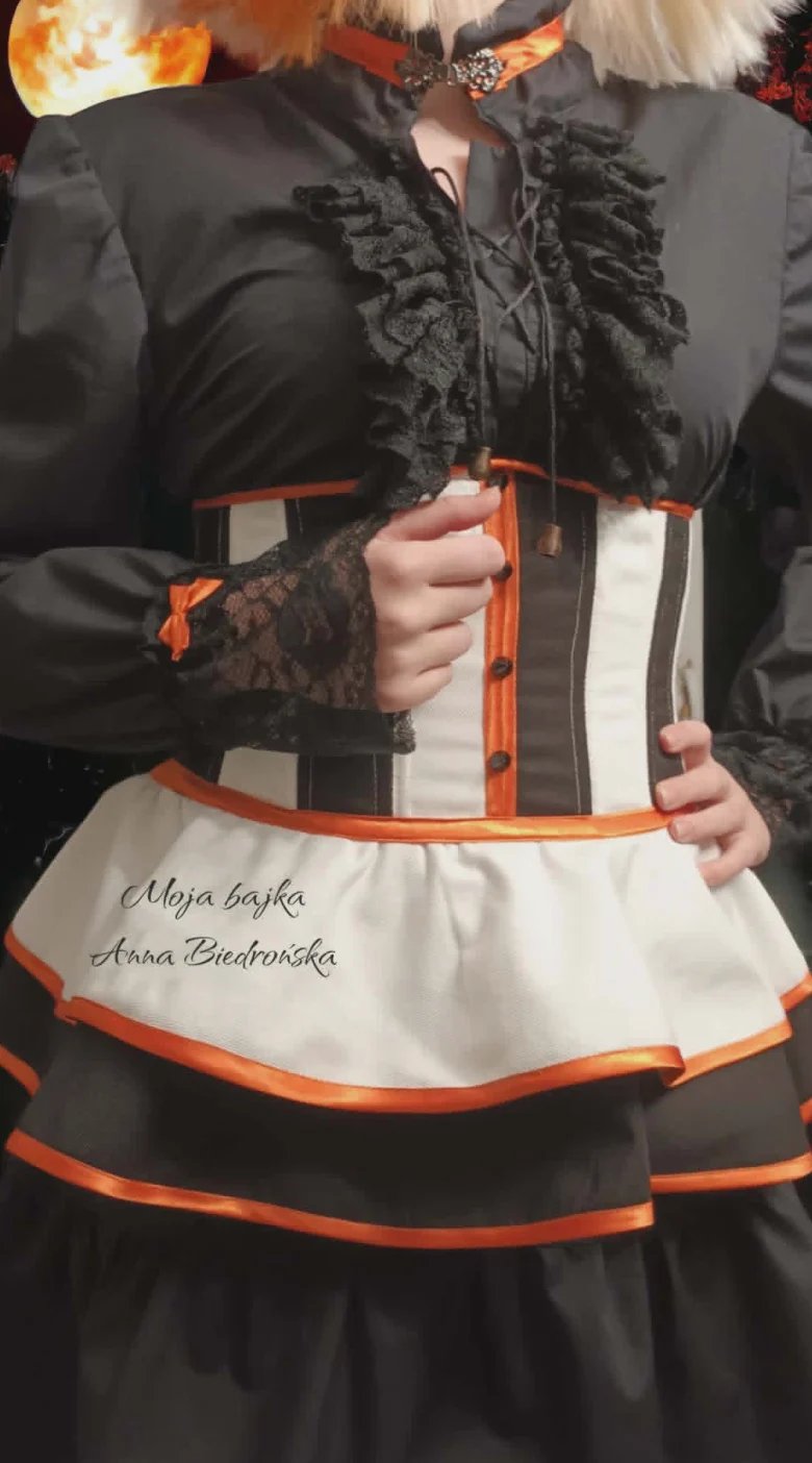 Person wearing traditional Polish folk costume with black embroidered vest, orange and white apron, holding ornamental staff