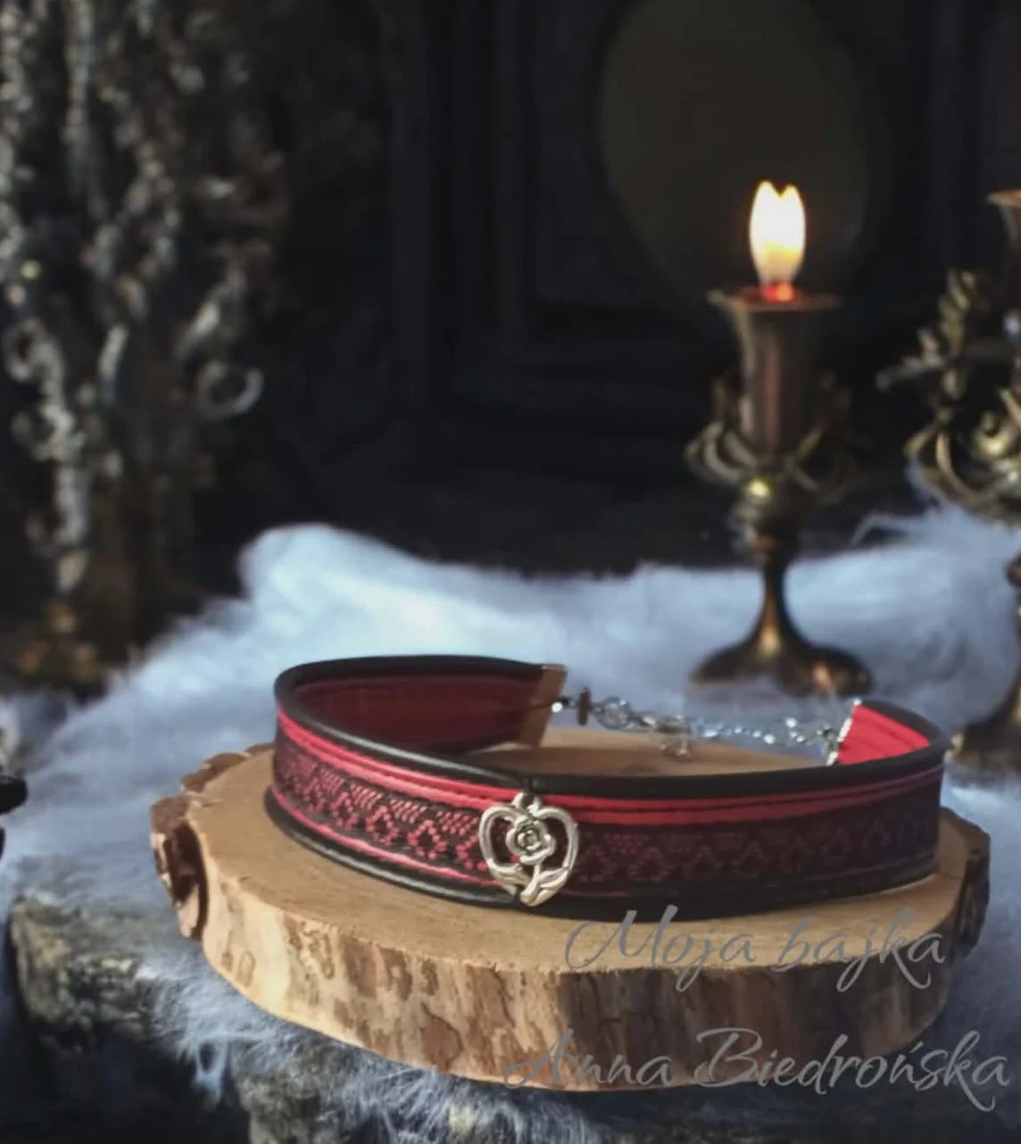 Red and black leather belt with ornate buckle displayed on wooden base with candles and gothic decor in background