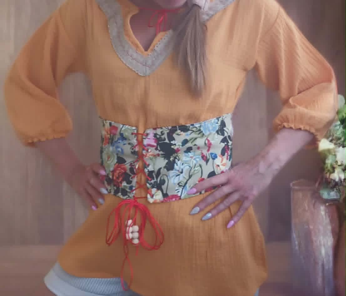 Person wearing an orange tunic with a floral belt and red drawstring detail, styled with a gray collar and long puffed sleeves.