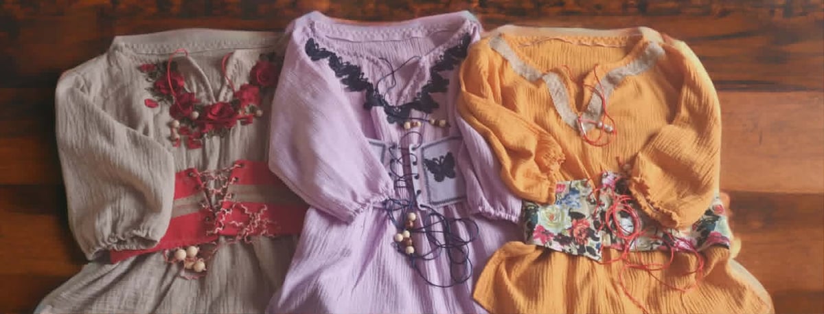 Three colorful childrens dresses displayed on a wooden floor: cream with red floral pattern, purple with dark trim, and yellow with floral accents