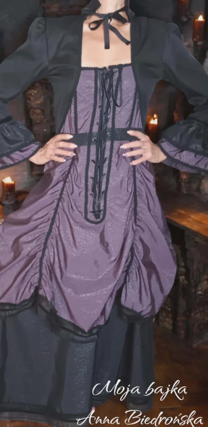 Woman wearing a dark blazer over a purple corset-style dress with black lacing and bow detail at neckline