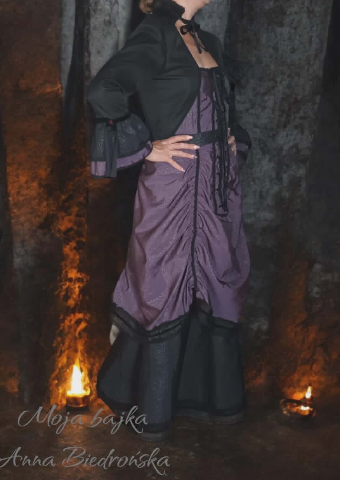 Woman in black jacket and purple dress standing between lit torches in a dark atmospheric setting