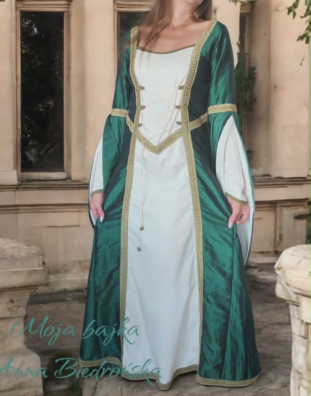 Woman wearing green and white medieval-style gown with gold trim and long sleeves, standing outdoors against a beige building.