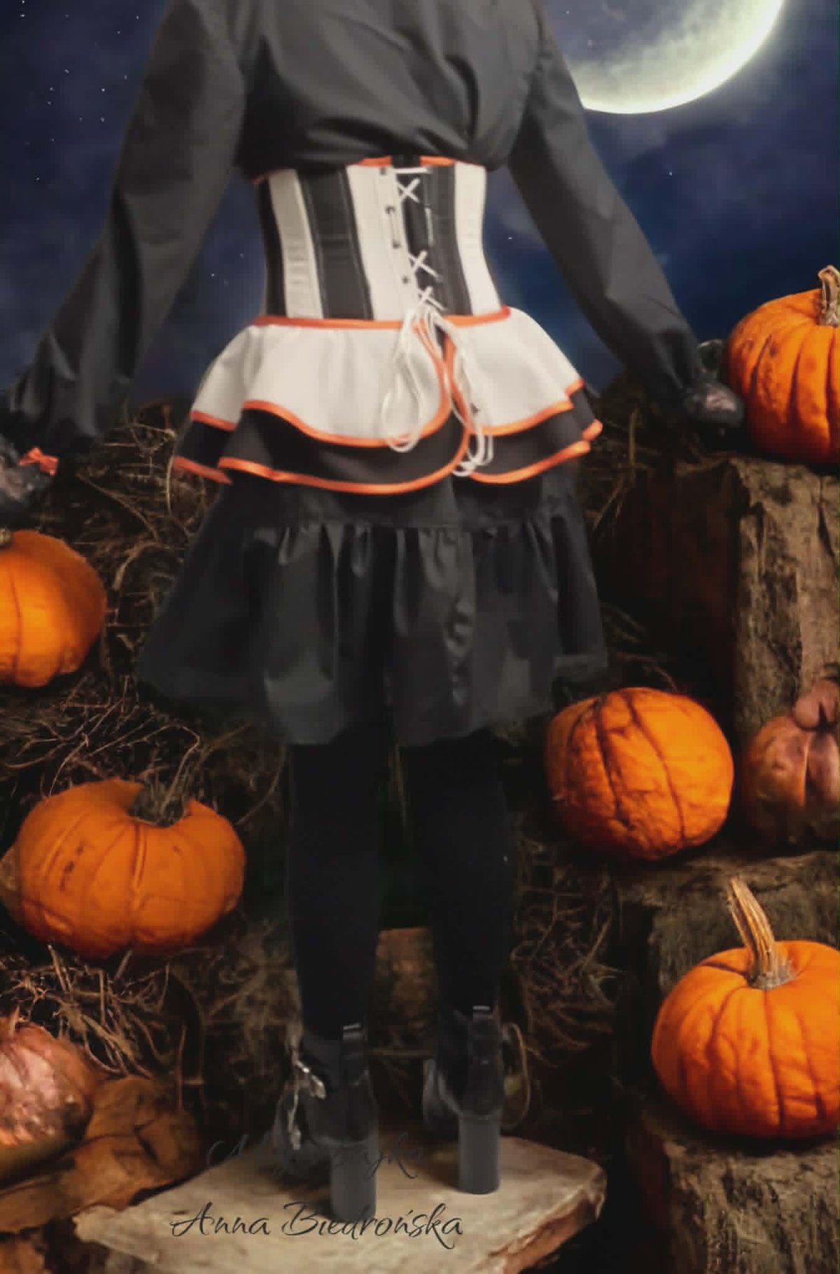 Person in Halloween witch costume with black dress and orange-trimmed corset standing in pumpkin patch under full moon at night