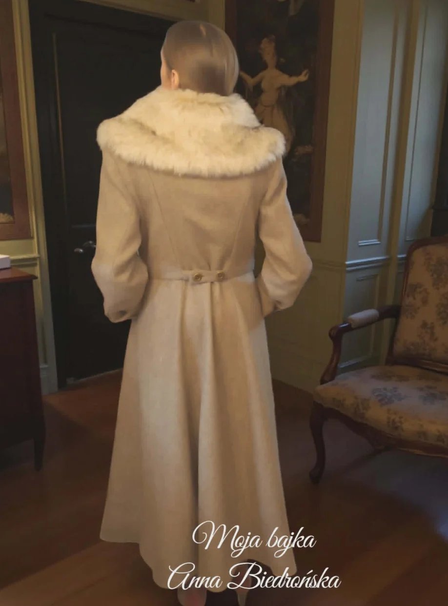 Woman wearing cream-colored vintage coat with fur collar viewed from behind in an elegant interior room