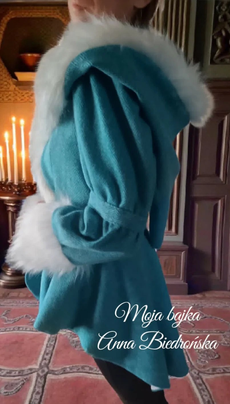 Child wearing a teal blue hooded coat with white fur trim, standing indoors near a candelabra and ornate furnishings