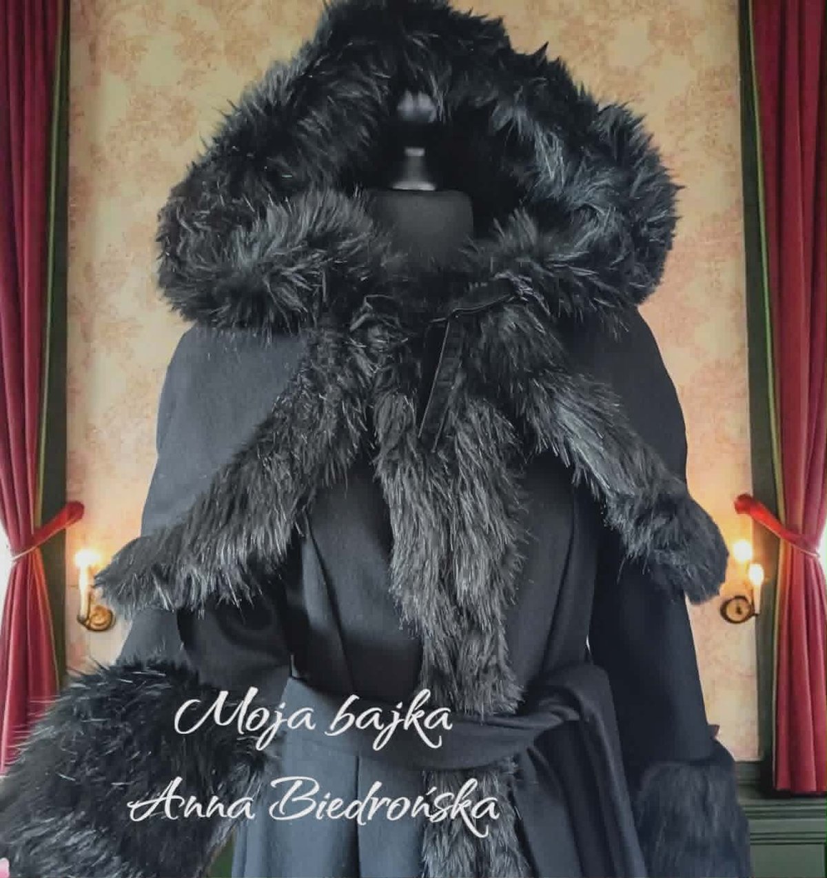 Elegant woman wearing black coat with large black fur collar and hood, posed against vintage theatrical backdrop with red curtains