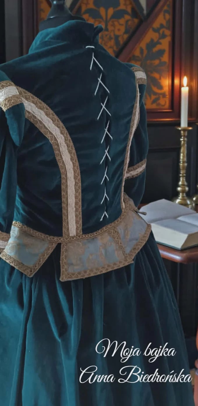 Back view of a deep teal medieval dress with gold embroidered trim and lace-up corset detail, displayed on a mannequin in a period room with candlelight