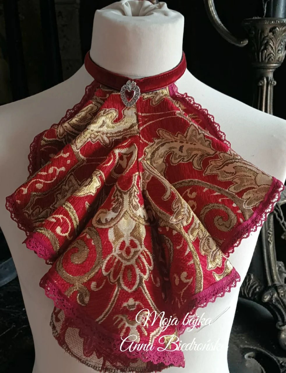 Ornate red and gold embroidered bow with lace trim displayed on a white dress form mannequin