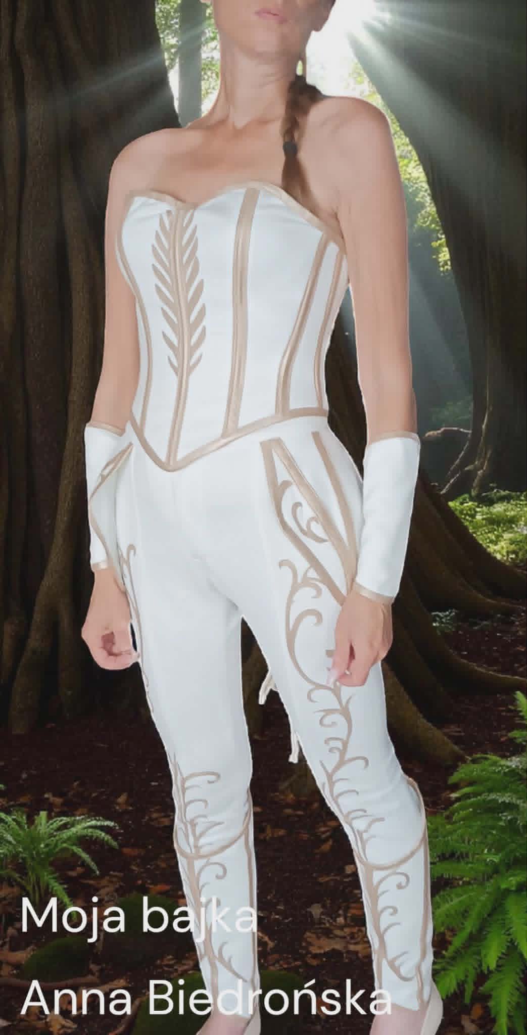 Woman in white strapless dance costume with tan leaf and scroll designs, wearing white arm covers, posing outdoors among trees