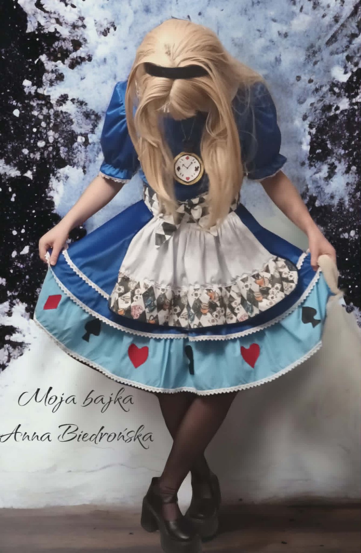 Person in blue Alice in Wonderland-inspired costume with blonde hair, holding skirt, wearing clock pendant against textured background