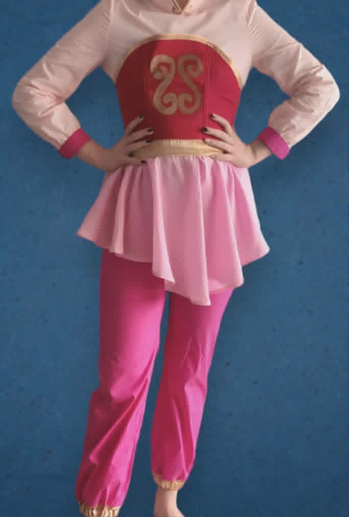 Person wearing a pink and red costume with ornamental chest piece, matching pink leggings, and a layered pink skirt against a blue background.
