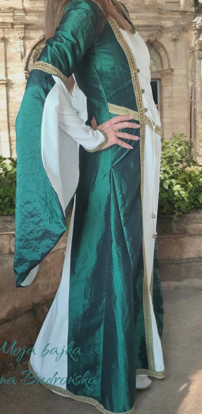 Woman wearing emerald green medieval-style gown with white underskirt and gold trim standing before stone church building