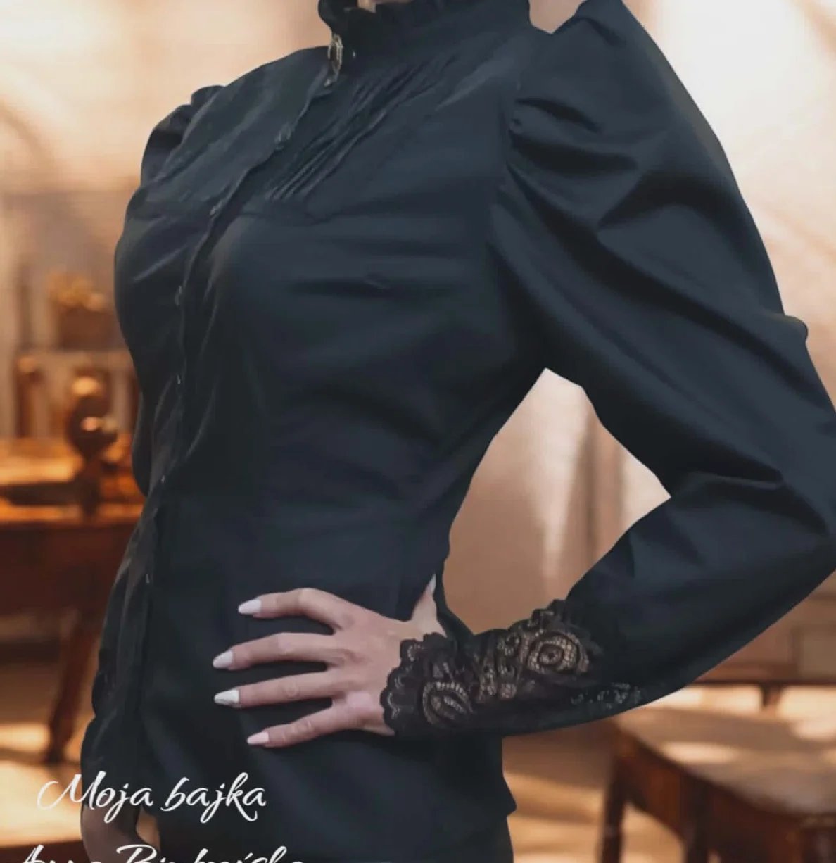 Woman wearing black dress with puffed sleeves and lace cuffs, hand on waist, posed indoors with warm lighting