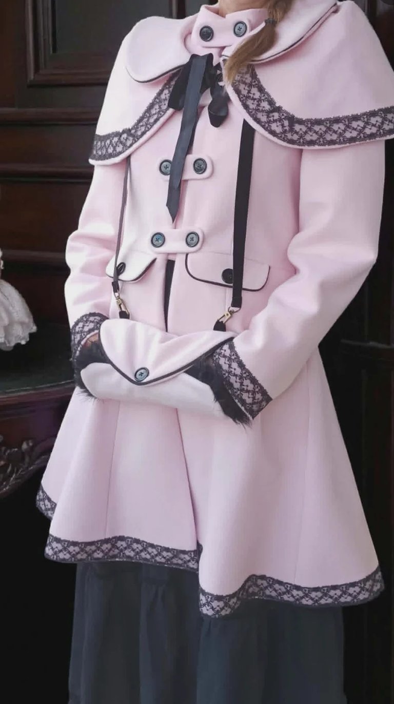 Person wearing a pink double-breasted coat with white collar, black lace trim, and matching cape with black ribbon tie
