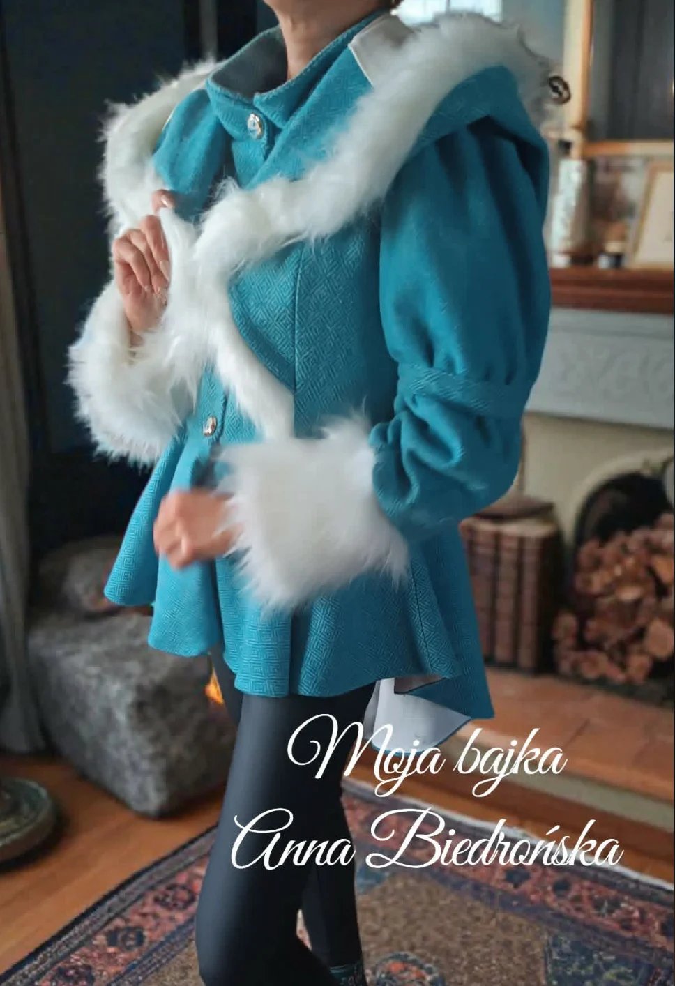 Person wearing turquoise coat with white fur collar and trim, black pants, standing indoors with winter home decor visible in background