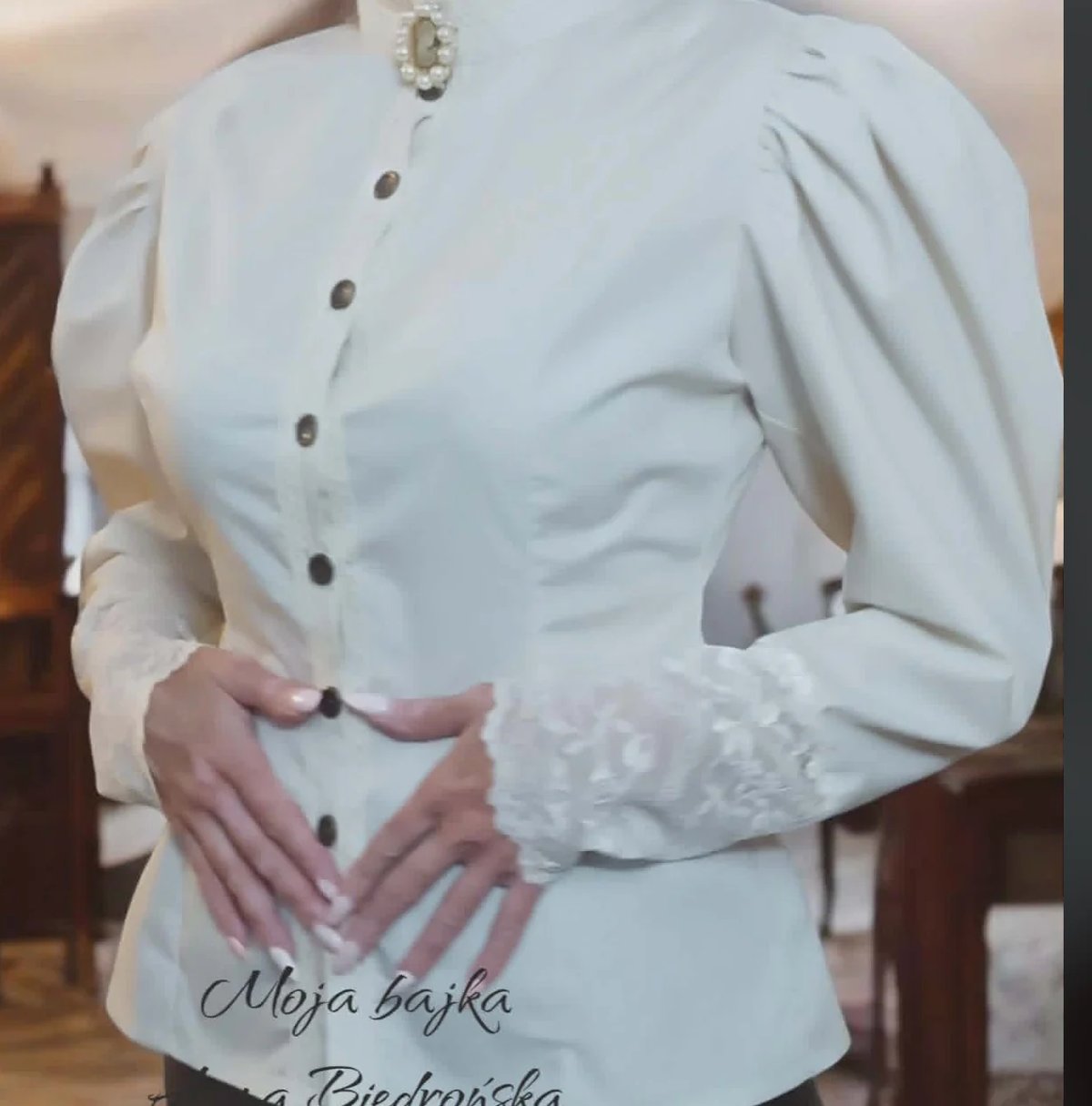 Woman wearing an elegant white blouse with puffed sleeves and pearl necklace, hands positioned at waist