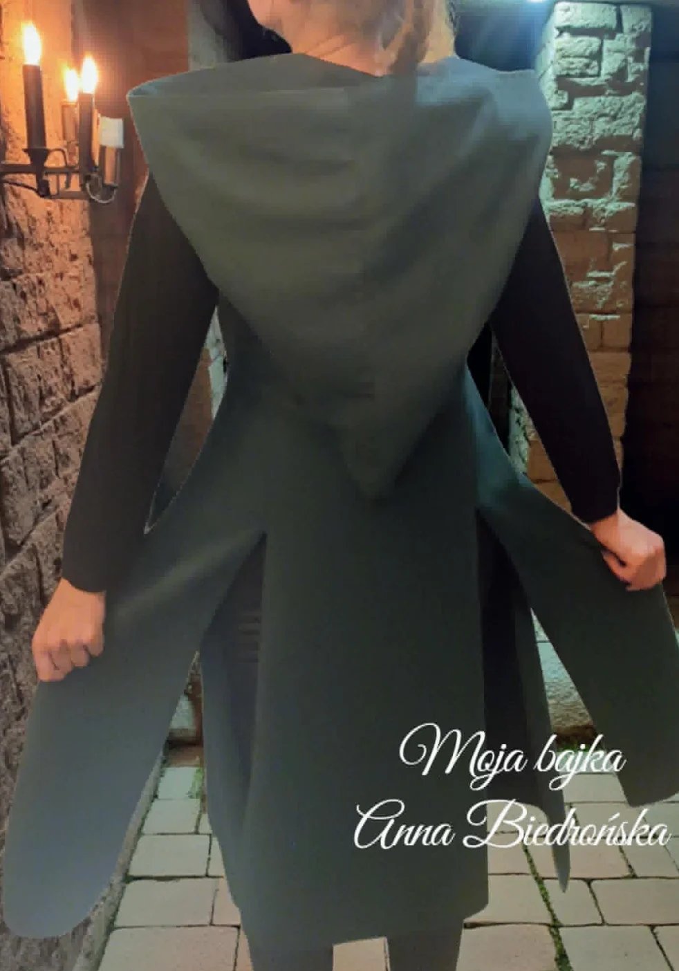 Person wearing a dark olive-colored hooded coat dress viewed from behind, standing on a brick pathway near lit candles and stone walls