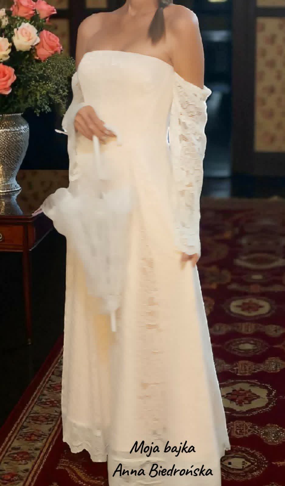 Pregnant woman in elegant off-shoulder white wedding dress standing indoors with floral arrangement in background