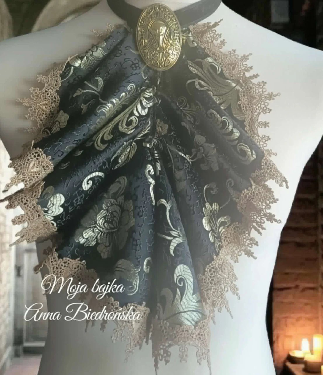 Ornate dark patterned fabric brooch with silver buckle and lace trim pinned to white fabric, with decorative text overlay