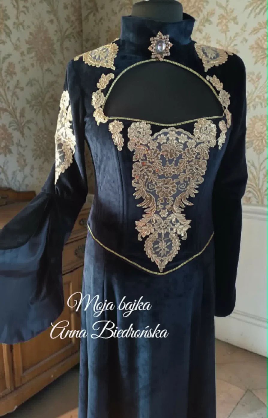 Black historical dress with ornate gold embroidery on shoulders and bodice, displayed on a mannequin with damask wallpaper background